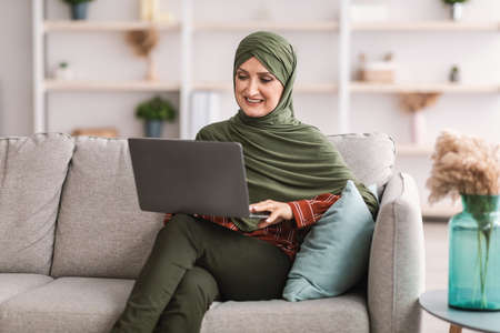 Cheerful Mature Muslim Female Using Laptop Sitting On Couch Indoorの写真素材
