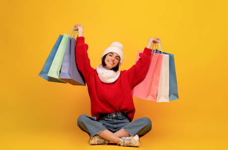 Happy young woman in winter clothes holding colorful shopping bags, sitting on yellow background
