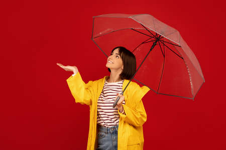 Rainfall concept. Happy young lady standing under umbrella and checking rain with outstretched hand, red backgroundの写真素材