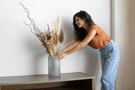 Modern minimalist home interior design. Young woman arranging dried plants in vase, creating cozy...
