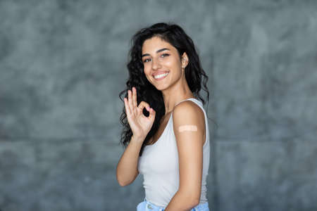 Young woman with plaster after virus vaccine injection gesturing okay over grey background