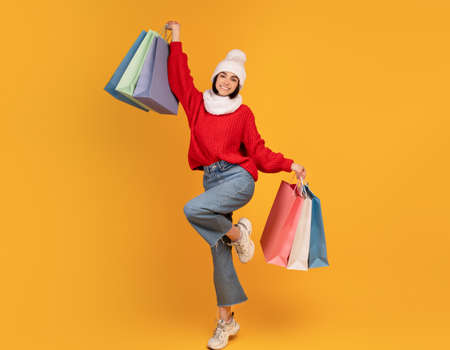 Excited winter lady holding colorful shopping bags and smiling to camera, posing over yellow...