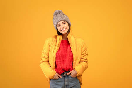 Happy lady in warm jacket, knitted sweater and hat holding hands in pockets and smiling to camera,...