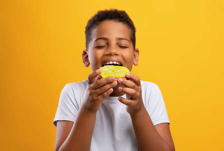 Black boy eating colorful donut, isolated on yellow backgroundの写真素材