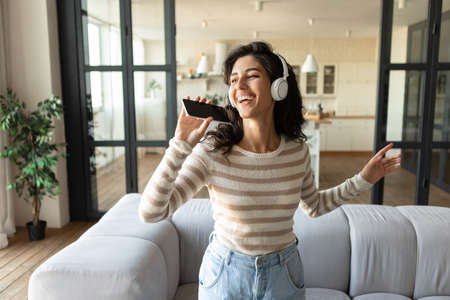 Pretty Caucasian woman in wireless headphones using new music application on smartphone, singing...