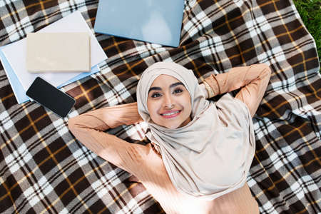 Glad cheerful attractive millennial arab middle eastern woman in hijab lies on blanket with booksの写真素材