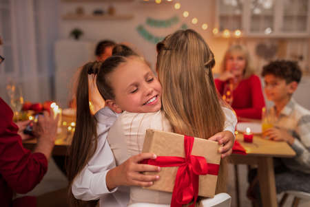Happy little girl hugging her mother, receiving gift box during festive dinner at home