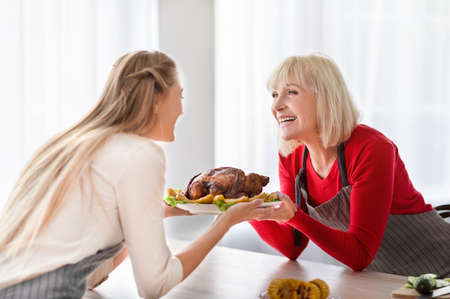 Happy senior woman holding Thanksgiving or Christmas turkey together with her young grown daughter at kitchenの写真素材