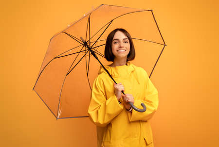 Autumn accessories. Pretty young woman holding transparent umbrella, smiling at camera over yellow backgroundの写真素材