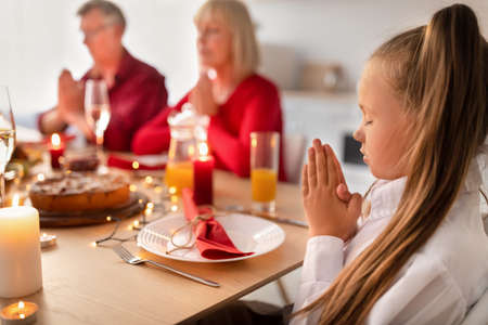 Little girl sitting at table, praying with grandparents before family dinner, celebrating holiday...
