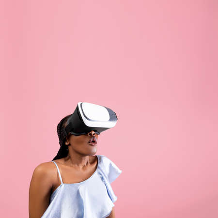 Cyberspace. Shocked African American woman wearing VR helmet, experiencing virtual reality on pink...