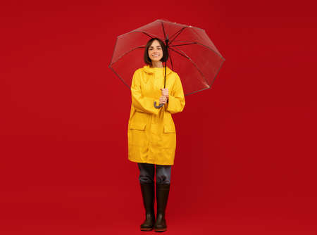 Autumn style. Studio shot of millennial lady in raincoat, standing under umbrella and posing over red backgroundの写真素材