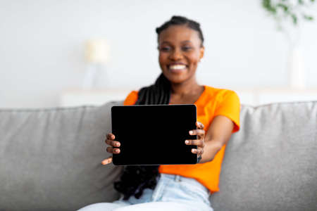 Cheery Afro woman showing tablet pc with blank screen, buying goods in internet store from home, mockupの写真素材