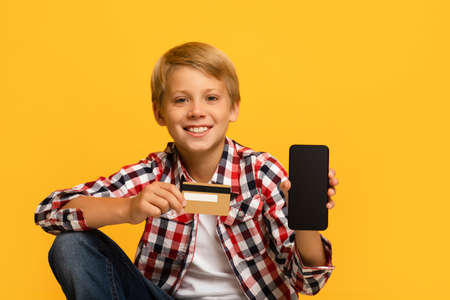 Cheerful handsome teen blond boy pupil show smartphone with blank screen and credit cardの写真素材
