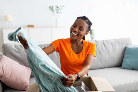 Satisfied buyer. Cheery black lady opening package after delivery, taking out new blouse, sitting on sofa at homeの写真素材