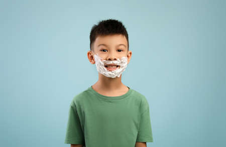 Little asian boy with shaving foam on face posing over blue background