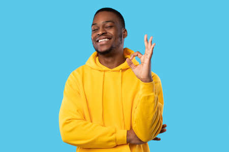 Cheerful Black Man In Yellow Hoodie Gesturing Okay, Blue Backgroundの写真素材