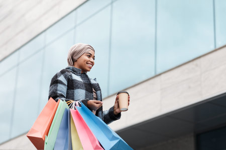 Glad happy pretty muslim black female in hijab and coat with bags of purchases and coffee takeaway, enjoying shoppingの写真素材