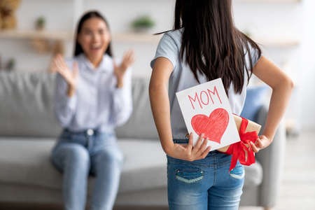 Mothers day gift. Girl congratulating excited mother with holiday, hiding greeting card and present box behind her backの写真素材