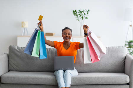 Happy black lady holding shopper bags and credit card, using laptop to place online order while sitting on sofa at homeの写真素材
