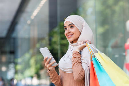 Smiling pretty millennial arabian lady in hijab enjoys online shopping with bags, surfing on phoneの写真素材