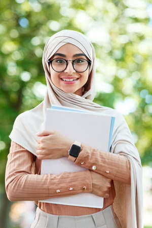 Cheerful glad cute young arab muslim female student in hijab and glasses holds books, ready for...