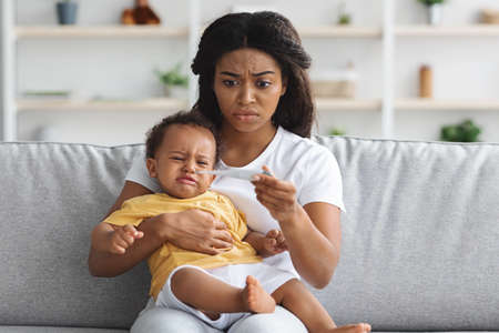 Worried Black Mother Checking Temperature Of Her Crying Infant Baby At Homeの写真素材