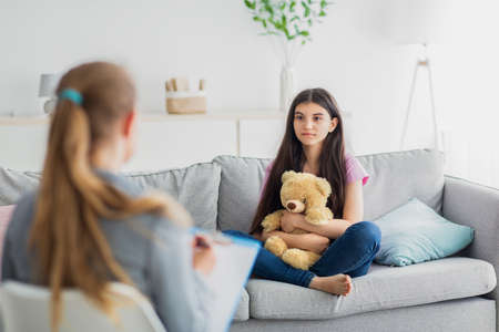 Foto de Indian teen girl having consultation with psychologist, sitting on couch with toy bear at psychotherapy clinic - Imagen libre de derechos