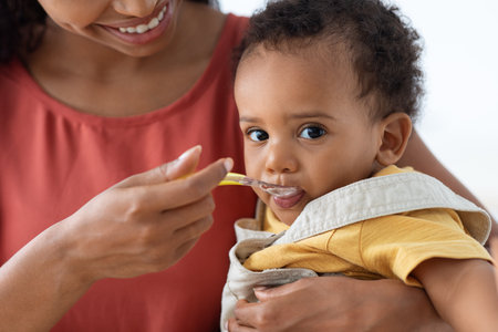 Happy Young Black Mother Spoon Feeding Her Cute Infant Baby Son, Closeupの写真素材
