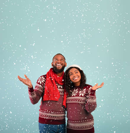 Xmas season. Joyful african american couple in warm winter clothes enjoying first snow, blue background with free spaceの写真素材
