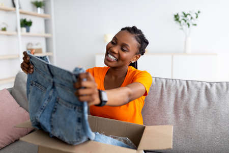 Ordering clothes online. Smiling black female customer opening parcel box, looking at new jeans on sofa at homeの写真素材