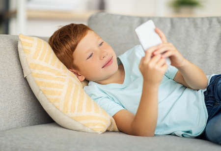 Happy boy laying on couch, using smartphoneの写真素材