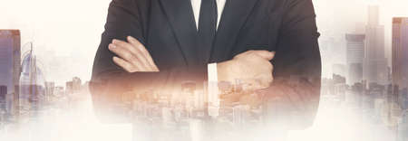 Cropped confident european young businessman in suit with crossed arms, double exposure with city skyscrapersの写真素材