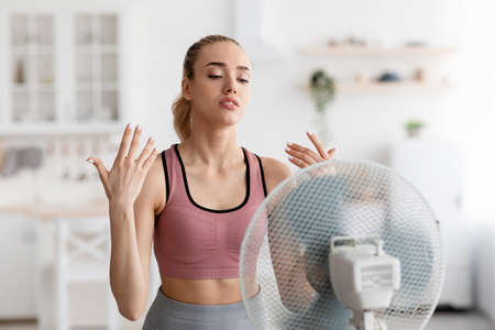 Tired sad european pretty millennial blonde lady stands in front of fan cooling down after workout...