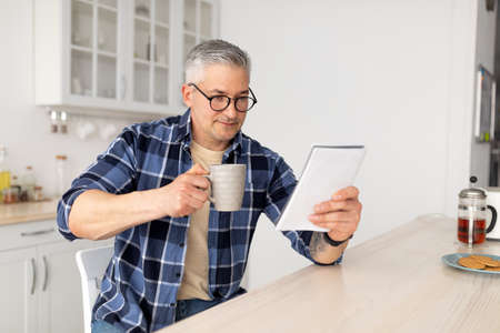 Morning routine. Senior man reading his notepad while sitting at kitchen table and drinking coffee, free spaceの写真素材