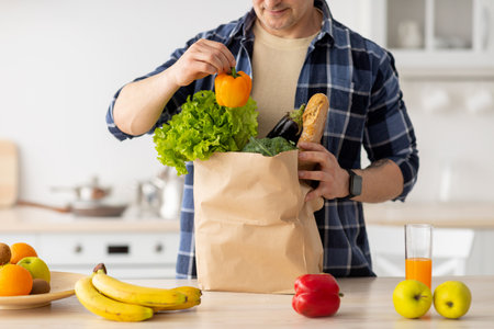 Mature man unpacking paper bag with fresh vegetables and fruits after grocery shopping, standing in kitchen, cropの写真素材