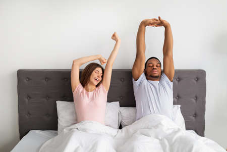 Multinational couple stretching on bed after waking up, feeling sleepy at homeの写真素材