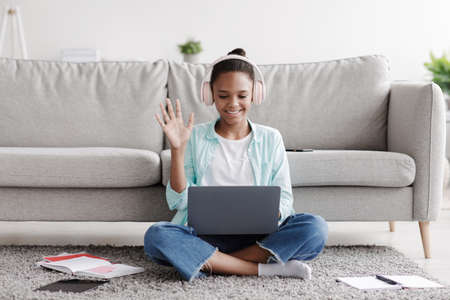 Cheerful teen black girl learns remotely, calls online and waves hand to laptop, sitting on floor at...