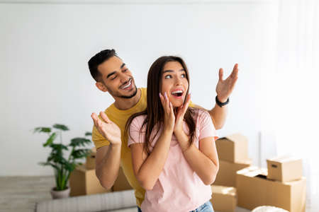 Excited Arab guy surprising his Caucasian girlfriend, moving to their own home together, standing in...