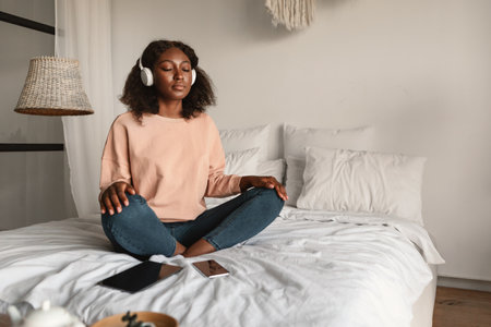 Black Woman Meditating Closing Eyes Wearing Headphones Relaxing In Bedroomの写真素材