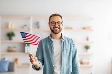 Handsome young guy holding American flag and smiling at camera indoors. Abroad education and student exchangeの写真素材