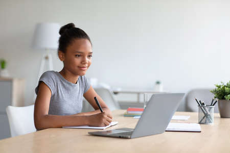 Smiling pretty smart teen african american girl student studying at home, watching online lesson on...