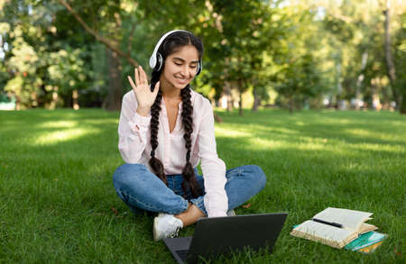 Remote learning. Excited indian female student using laptop, having online lesson and waving to webcam, sitting outdoorsの写真素材
