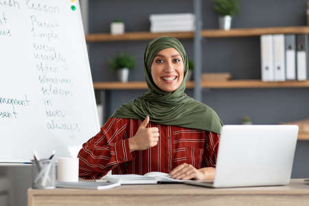 Islamic teacher in headscarf sitting at desk showing thumbs upの写真素材