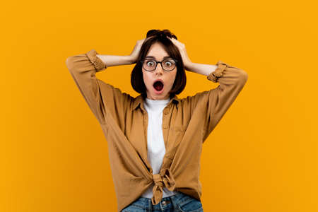 Unbelievable news. Shocked woman touching her head in shock over yellow studio background, cannot...