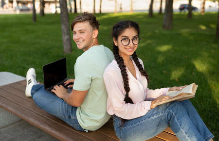 Modern students using laptop and book, learning outdoors, sitting back to back on bench in park outsideの写真素材