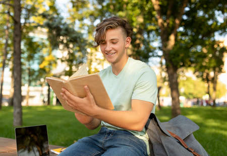 Education concept. Teenage student guy reading book outdoors and preparing for exams, sitting in...