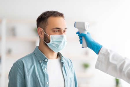 Doctor in rubber gloves measuring young guys temperature with infrared thermometer indoorsの写真素材