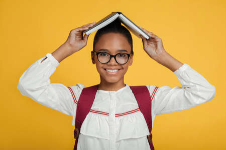 Smiling smart young african american female pupils in glasses with backpack holding book on her headの写真素材