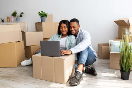 Happy african american spouses having video call via laptop, sitting among carton boxes in new...